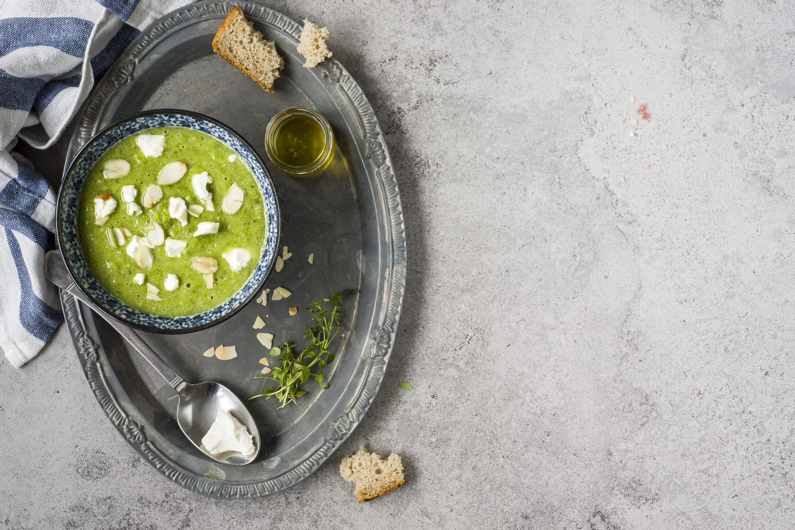 Zielona zupa krem podana w niebieskiej misce, ozdobiona płatkami migdałów i kozim serem, z dodatkiem świeżych ziół i oliwy z oliwek - idealny przepis na zdrowy i lekki obiad.